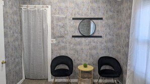 Habitació amb cortina de dutxa, dues cadires negres, una taula rodona petita amb una planta verda i un mirall fixat a la paret.