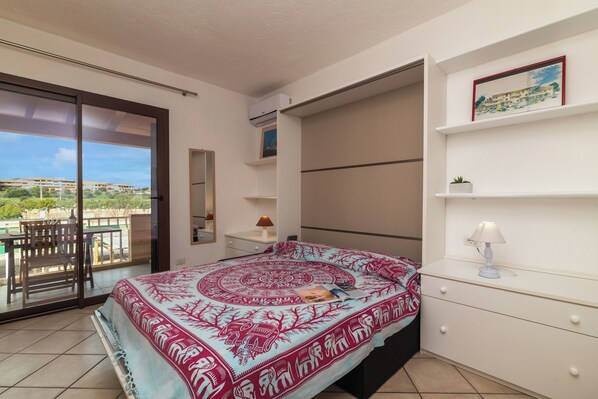 A bedroom with a bed, a nightstand, a lamp, a sliding door to a balcony, and a view of buildings.