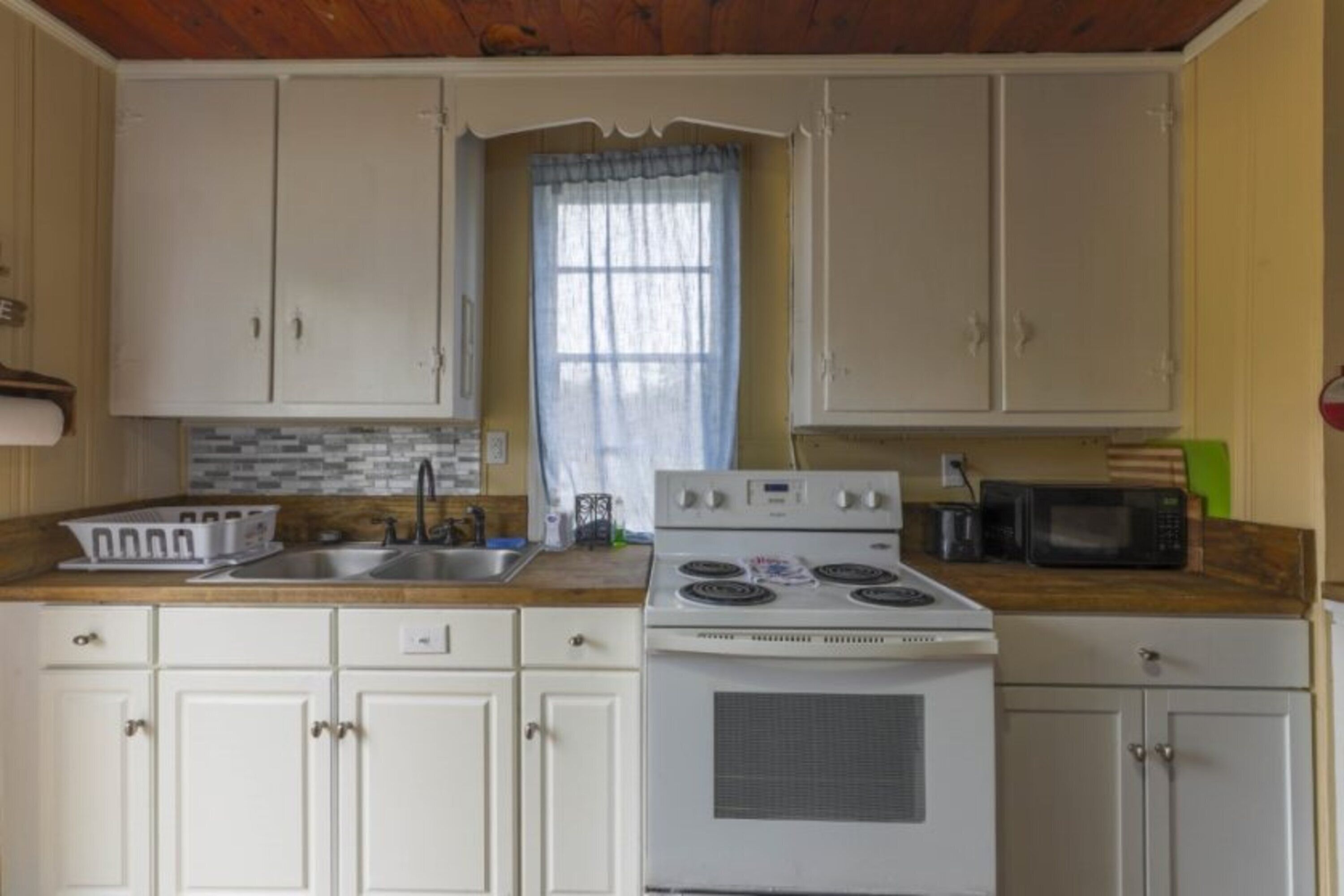 A kitchen with white cabinets, a stove, a microwave, and a window with a curtain.