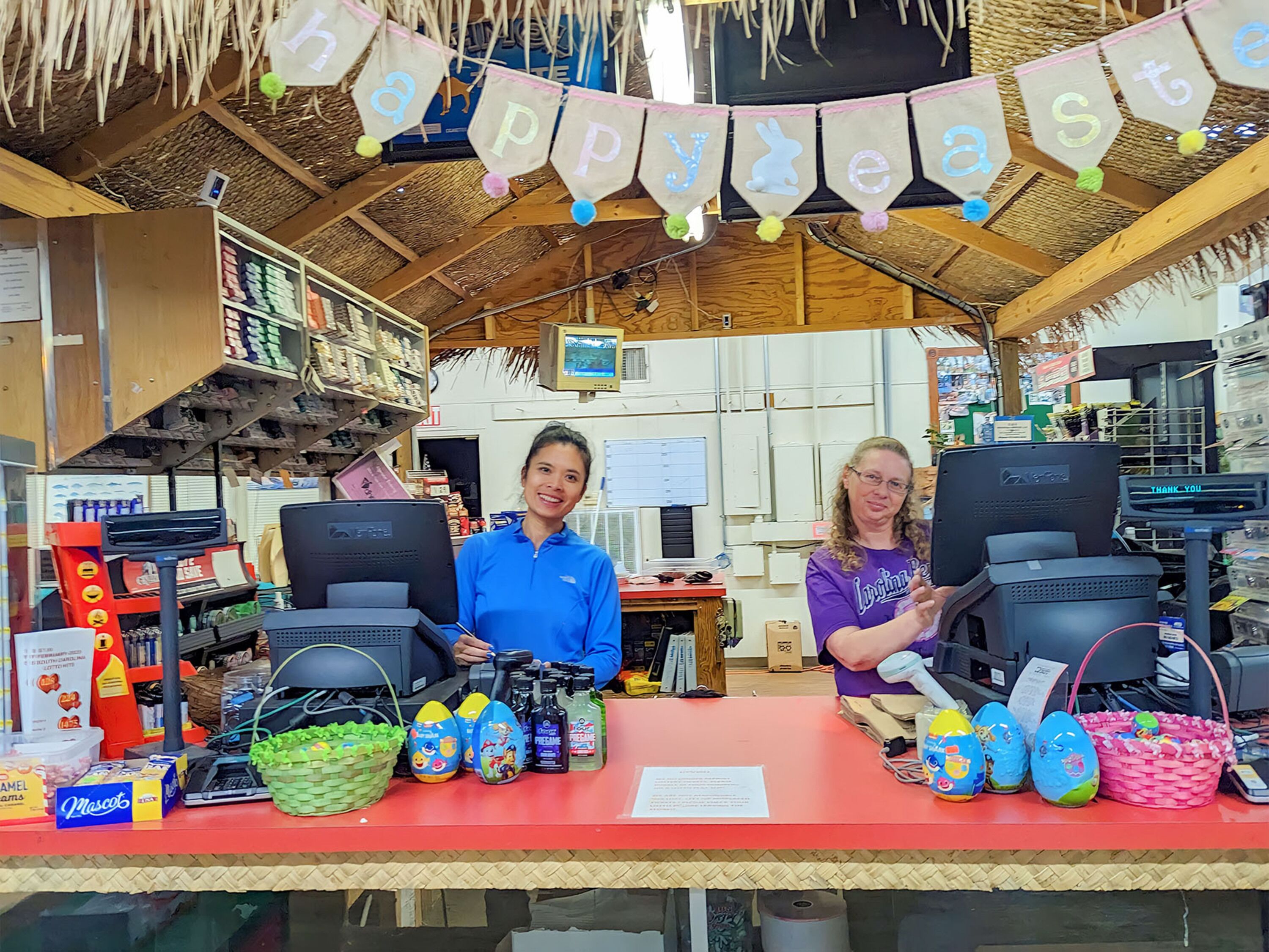 Una pequeña tienda con dos personas atendiendo, varios artículos a la vista y un cartel que dice 'Feliz Pascua'.