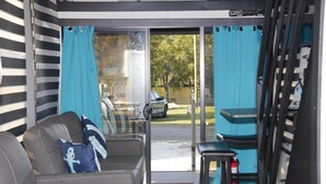 Une pièce moderne avec un canapé gris, une petite table et une vue sur un lac et une voiture garée, visible à travers de grandes portes vitrées.