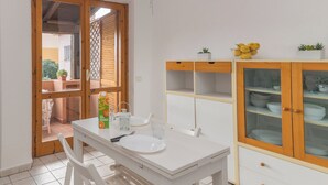 A dining area with a white table set for two, a wooden cabinet with glass doors, and a view through a door to an outdoor patio. - Piazzetta Bilo Brezza Marina (Loiri Porto San Paolo)
