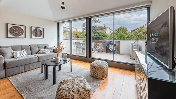 A modern living room with a grey sofa, a coffee table, and a TV. Large windows offer a view of the outside.