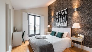 A modern bedroom with a large bed, a view through a sliding door, a wall-mounted artwork, and a nightstand with a lamp.