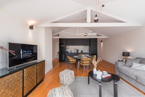 A modern living room with a flat-screen TV, a grey sofa, a dining area with a table and chairs, and a kitchen in the background. - Modern loft in the heart of Porto (Porto)