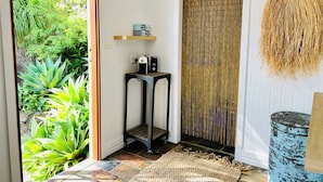A room with a view of lush greenery through a glass door, a bamboo screen, a small table with a kettle and a speaker, and a blue-painted barrel. - Surf Break On Bruxner - Korora (Pet Friendly) (Korora)