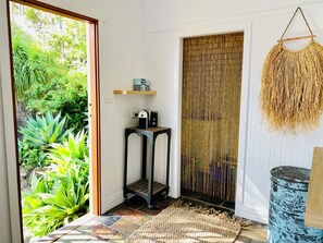 Une pièce donnant sur une végétation luxuriante grâce à une porte vitrée, un paravent en bambou, une petite table avec une bouilloire et un haut-parleur, et un tonneau peint en bleu.