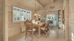 Un interno in legno con zona pranzo, sedie in vimini e un tavolo, ampie finestre e una cucina sullo sfondo.