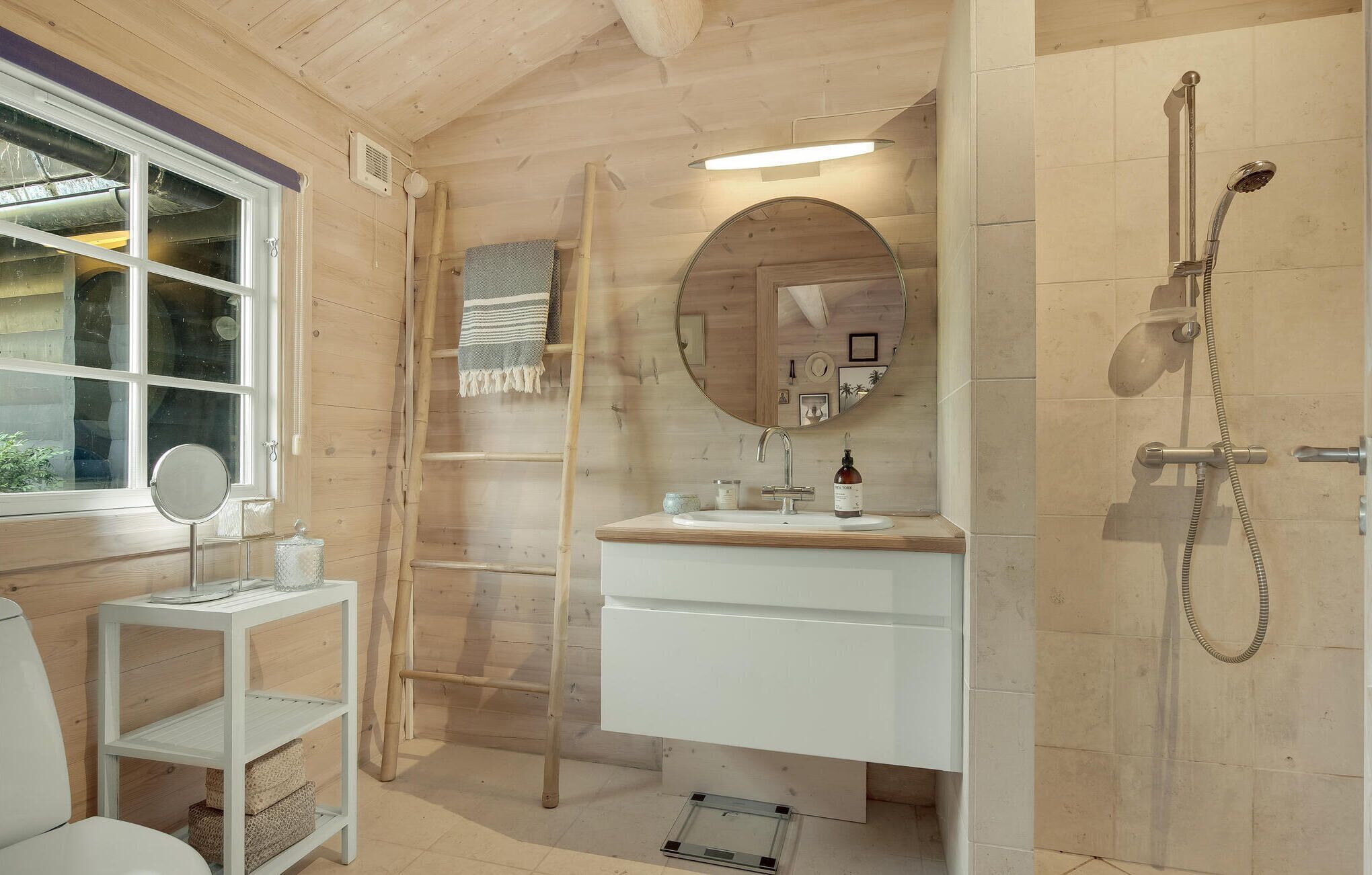 A bathroom with a wooden interior, a round mirror, a sink with a bottle, and a shower.
