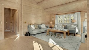 Accogliente interno di una baita in legno con un divano blu chiaro, un tavolino in legno e un'ampia finestra con tende.