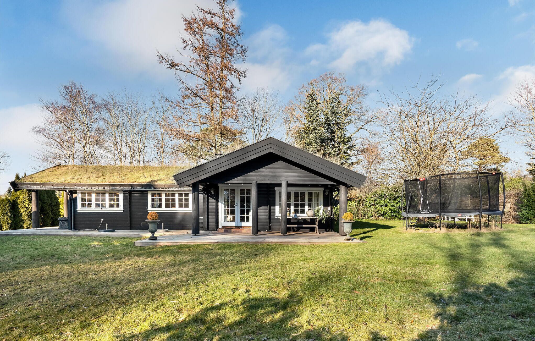 Une maison moderne dotée d’une grande véranda, d’un espace de détente extérieur abrité et d’un trampoline dans le jardin.