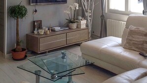 En stue med et sofabord i glas, en beige sofa, et træskab med pynteting og en potteplante.