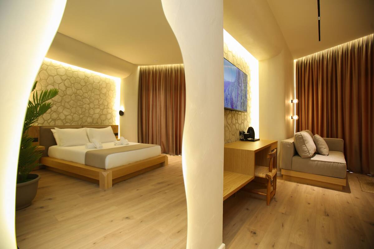 A modern hotel room with a wooden floor, a bed, a sofa, a desk, and a TV.