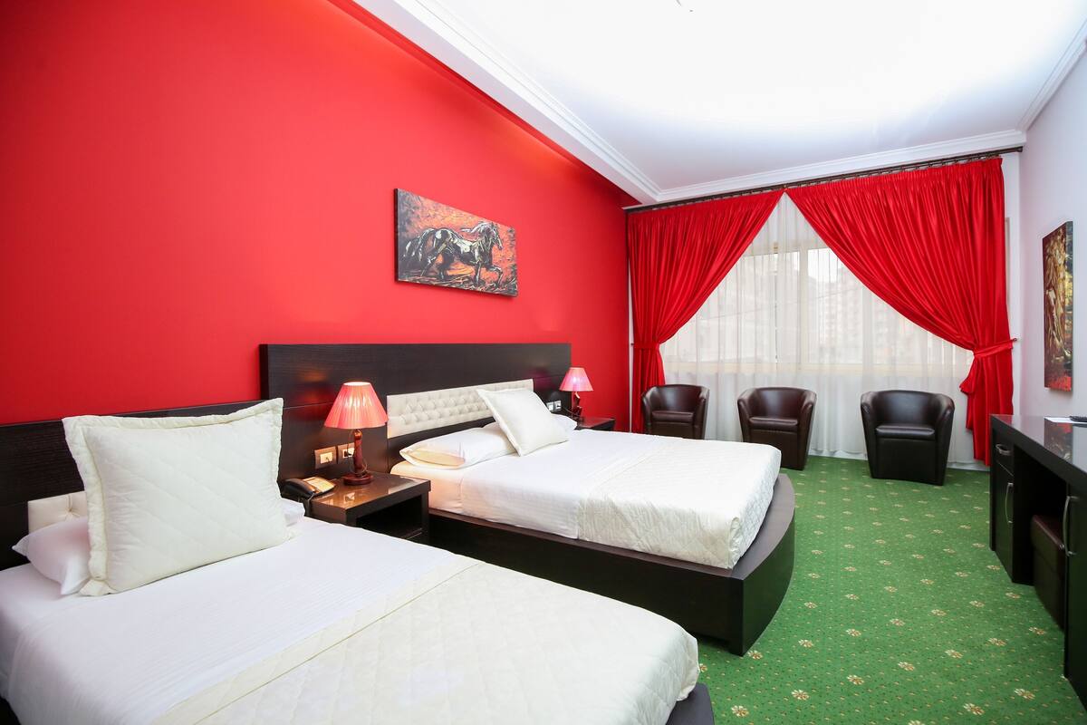 A hotel room with two beds, a red wall, a painting, a window with red curtains, and a green carpet.