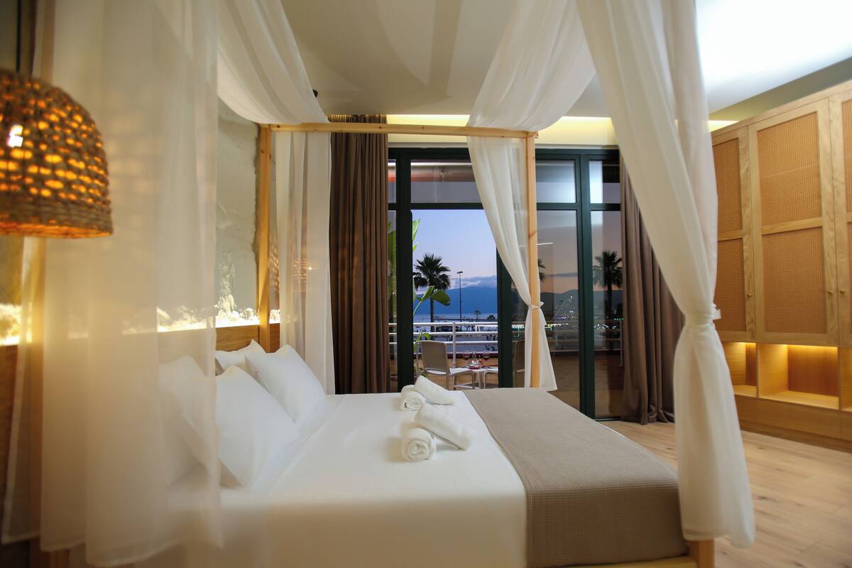 A hotel room with a bed, white curtains, a view of palm trees, and a balcony.