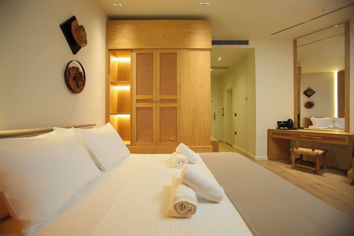 A hotel room with a neatly made bed, a wooden wardrobe, a desk with a chair, and a large mirror.
