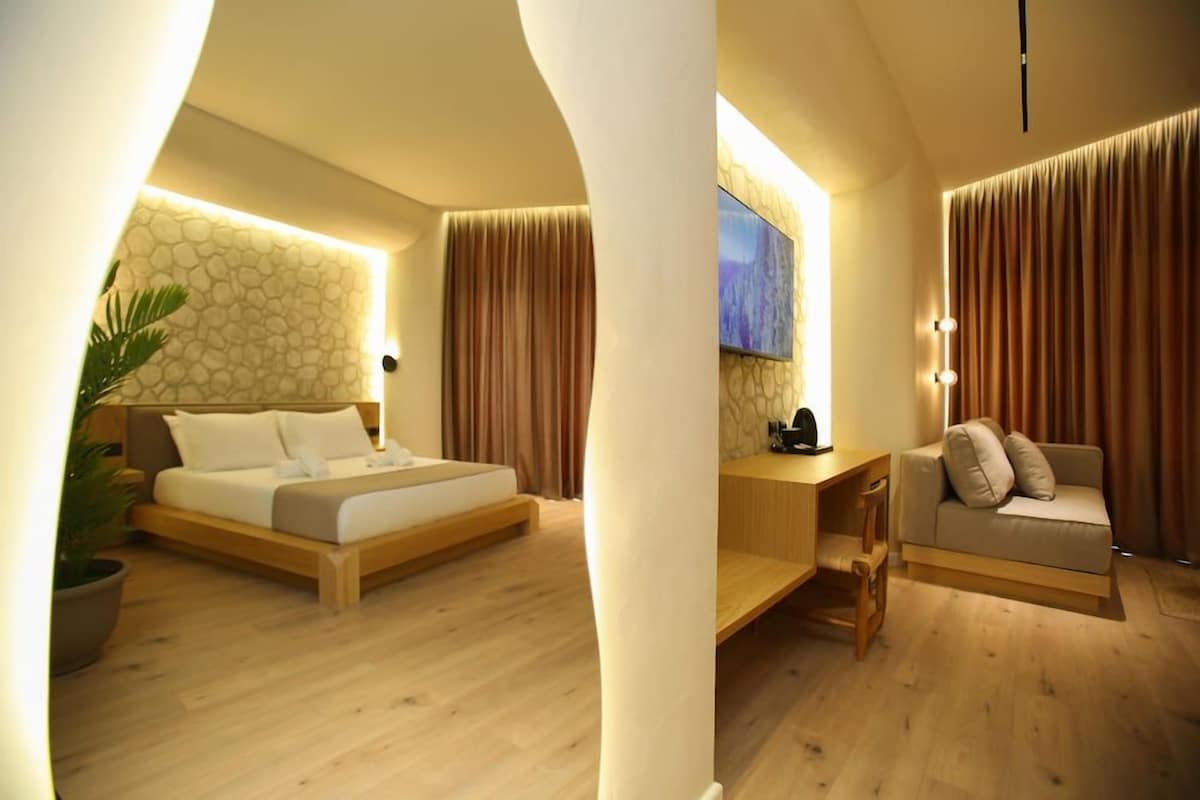 A modern hotel room with a wooden bed, a sofa, a desk, and a large window with curtains.