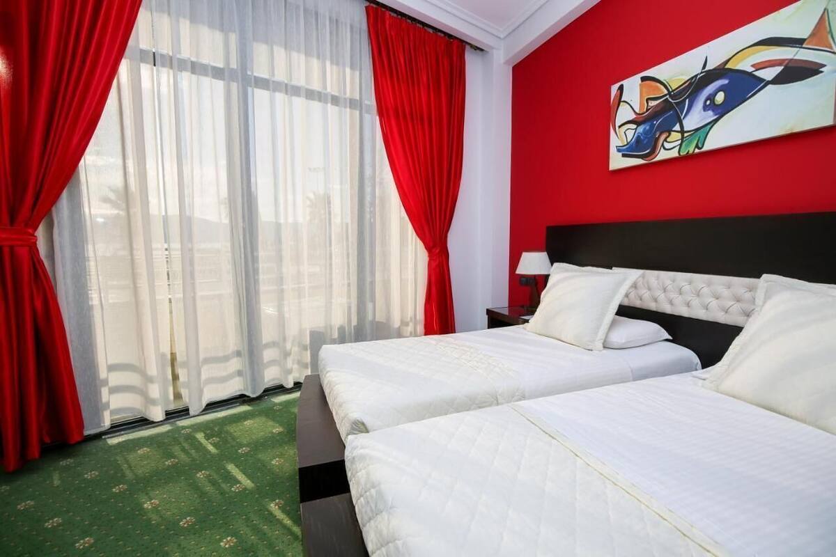 A hotel room with two beds, a red wall, and a window with sheer curtains.