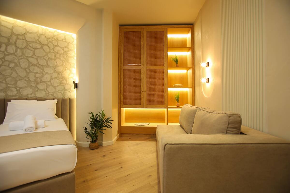 A hotel room with a bed, a sofa, a wooden cabinet with shelves, and a potted plant.
