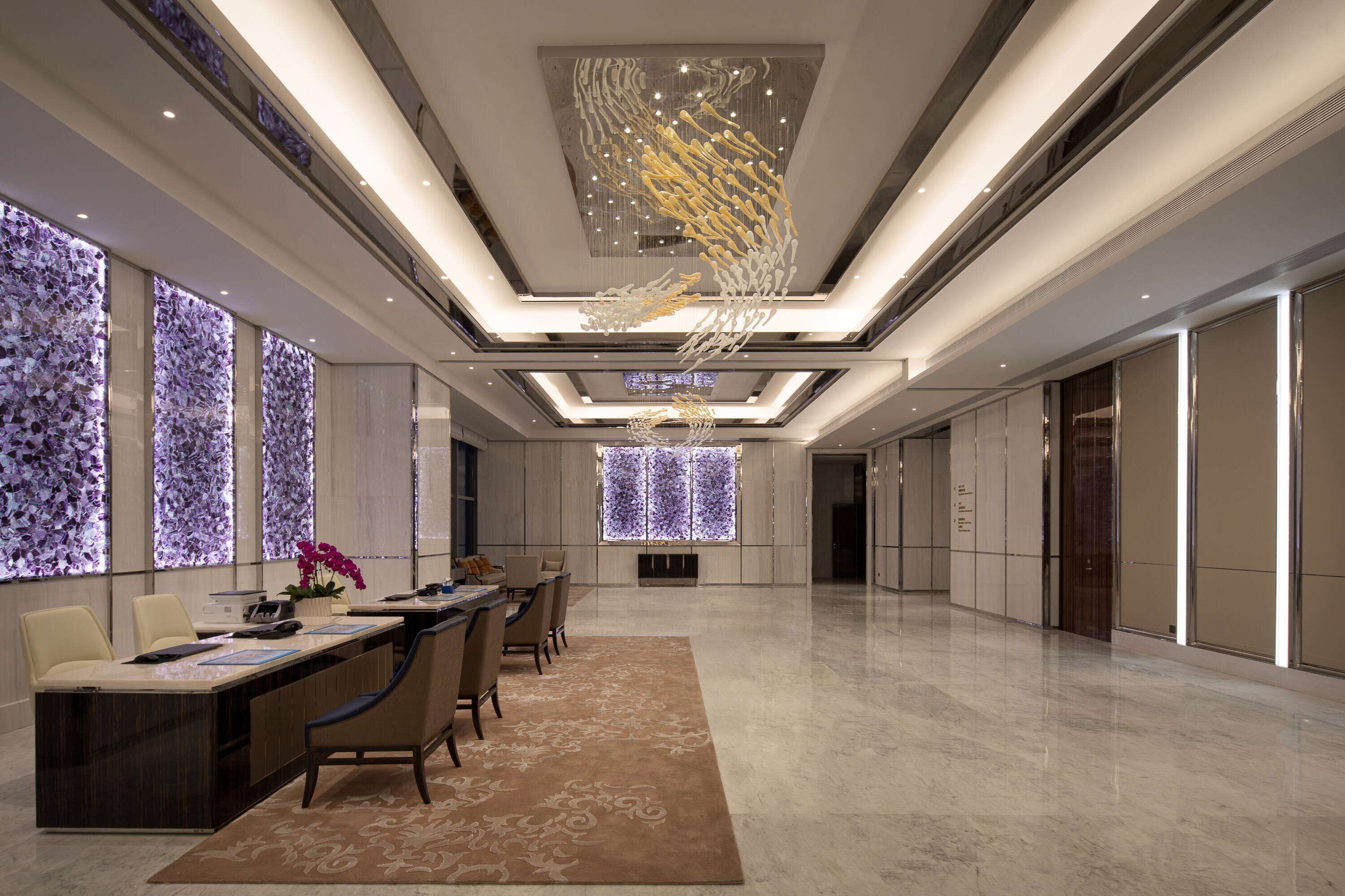 A spacious hotel lobby with a long reception desk, comfortable seating, and a modern chandelier.