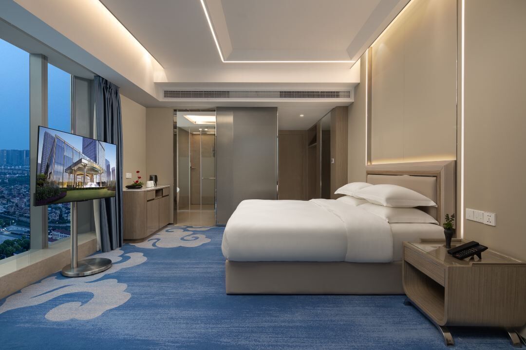 A modern hotel room with a large bed, a TV on a stand, a desk, and a view of the city.