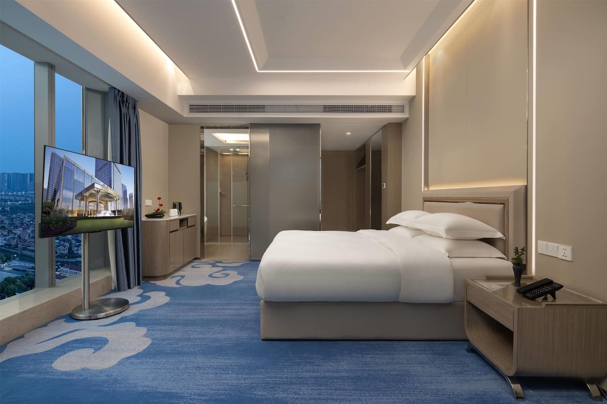 Ein modernes Hotelzimmer mit einem großen Bett, einem Fernseher auf einem Schrank, einem Nachttisch und Blick auf die Stadt durch große Fenster.