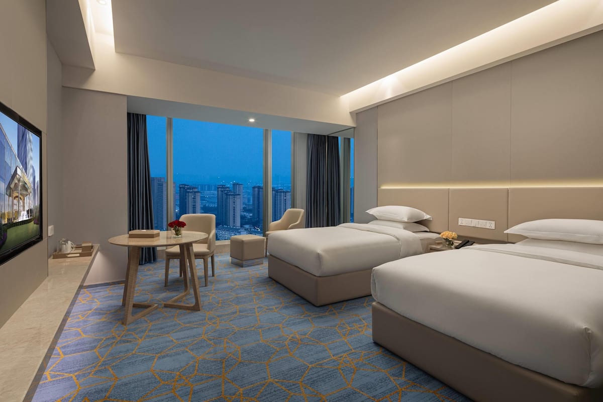 Ein Hotelzimmer mit zwei Betten, einer Sitzecke mit Tisch und einem großen Fenster mit Blick auf die Stadt.