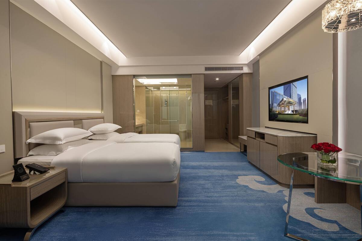 Ein modernes Hotelzimmer mit einem großen Bett, einem Fernseher, einem Glastisch und einem durch eine Glastür einsehbaren Badezimmer.