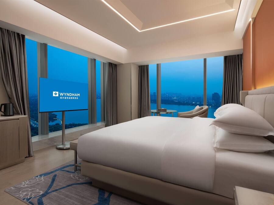 A modern hotel room with a large bed, a desk, and a TV screen displaying the Wyndham logo.