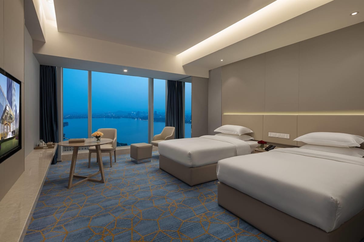 Ein Hotelzimmer mit zwei Betten, einem kleinen Tisch mit Blumenvase, einem Sessel und Blick auf die Stadt und das Wasser.