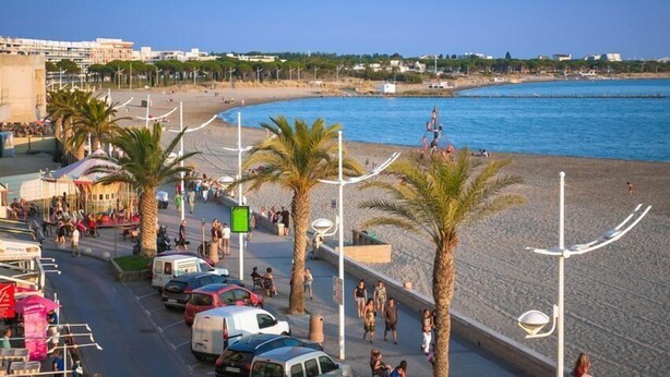Un paseo marítimo en la playa, con coches aparcados, palmeras y una vista despejada del mar.