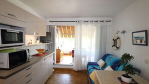 Cocina compacta con microondas, estufa y armarios. Cuenta con comedor con mesa y sillas, y una cama con colcha colorida. Hay una planta en maceta sobre la mesita de noche.
