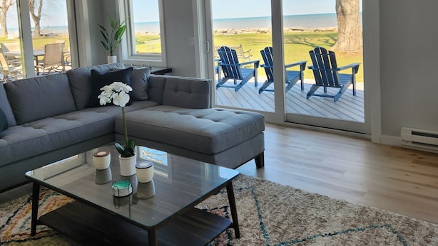 A modern living room with a large gray sectional sofa, a glass coffee table, and a ceiling fan. There are large windows offering a view of the outdoors, including a deck with chairs. - Tranquil Lakefront home. Premium accommodations, newly remodeled (Hamlin)