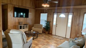 Ein gemütliches Wohnzimmer mit Holzwänden, einem Flachbildfernseher, einem Deckenventilator, einem beigen Sofa, einem weißen Sessel, einem Couchtisch und einer Topfpflanze.