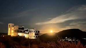 A modern building with multiple levels and balconies, situated on a hill with a sunset in the background.