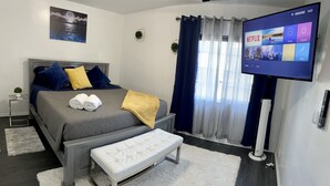 A modern hotel room with a bed, a bench, a flat-screen TV displaying the Netflix app, and a view of the outdoors through sheer curtains.