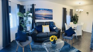 A modern living room with a black leather sofa, two armchairs, a glass coffee table, and a painting of a sunset over the ocean.