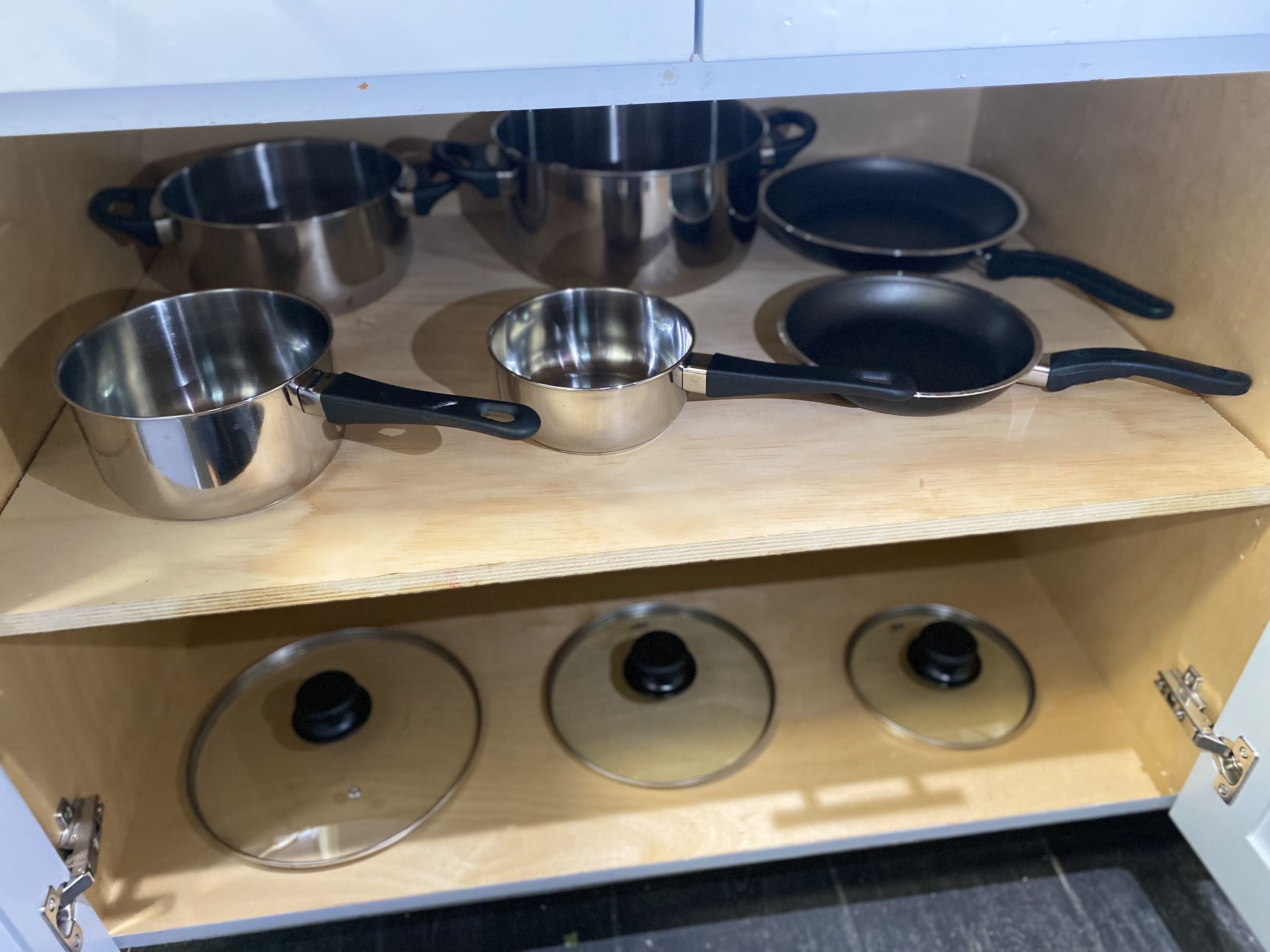 A kitchen cabinet with stainless steel cookware, including pots, pans, and lids.