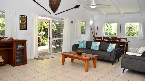 A spacious living area with a sofa, coffee table, and a dining table. There is a ceiling fan, a decorative wall piece, and a view of greenery outside. - Tiare Taporo (Matavera District)