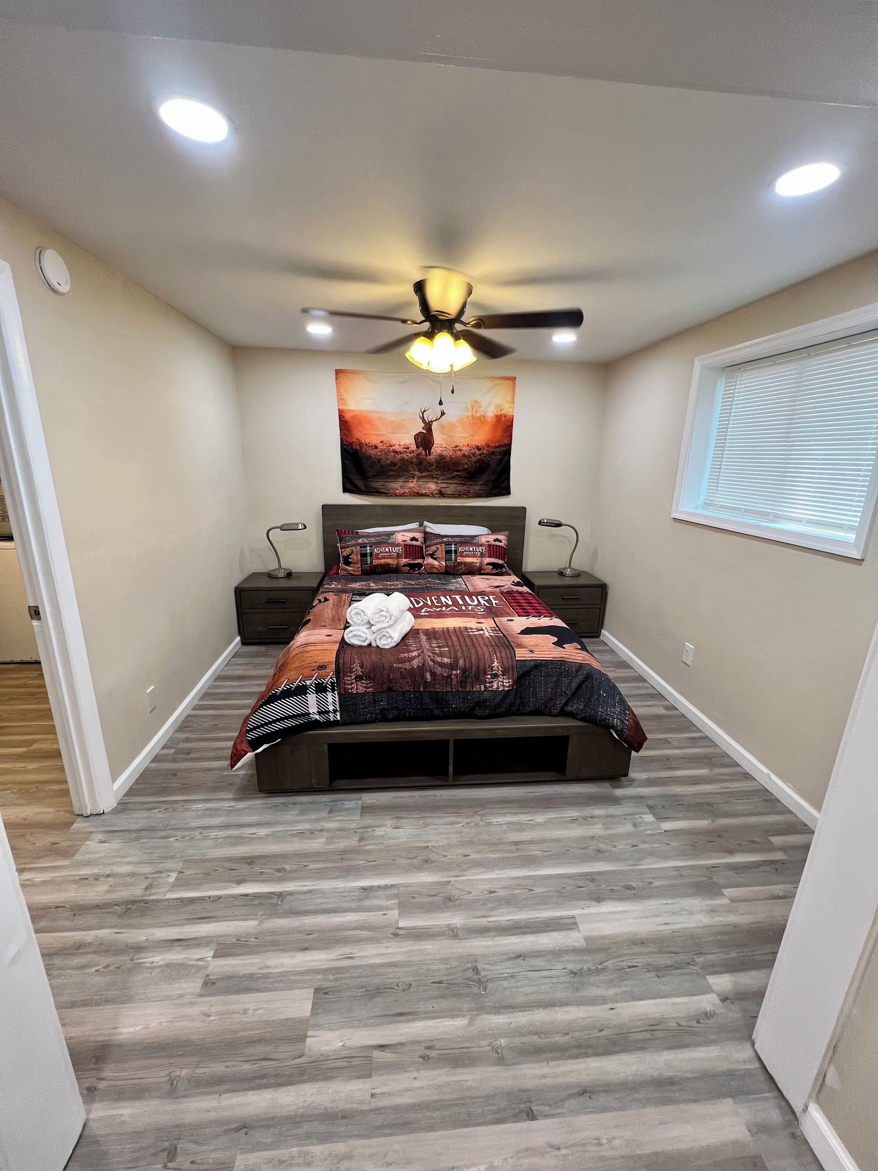 Une chambre à coucher comprenant un lit, un ventilateur de plafond, une lampe fixée au mur, une fenêtre avec des stores et une tapisserie murale.