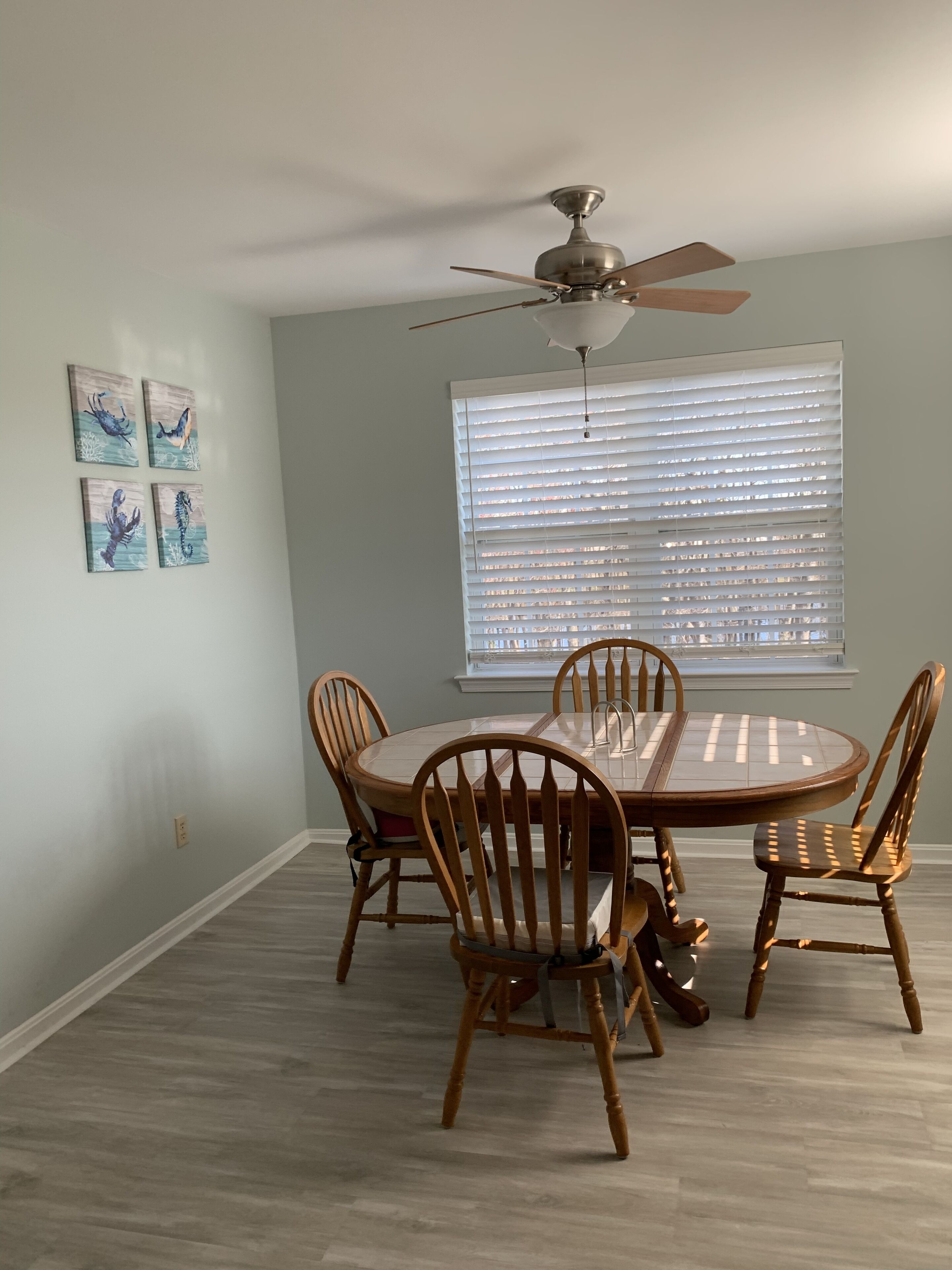 A dining area with a round table and four wooden chairs, a ceiling fan, and a window with blinds.