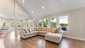 A spacious living room with a large sectional sofa, wooden flooring, and large windows offering a view of greenery.