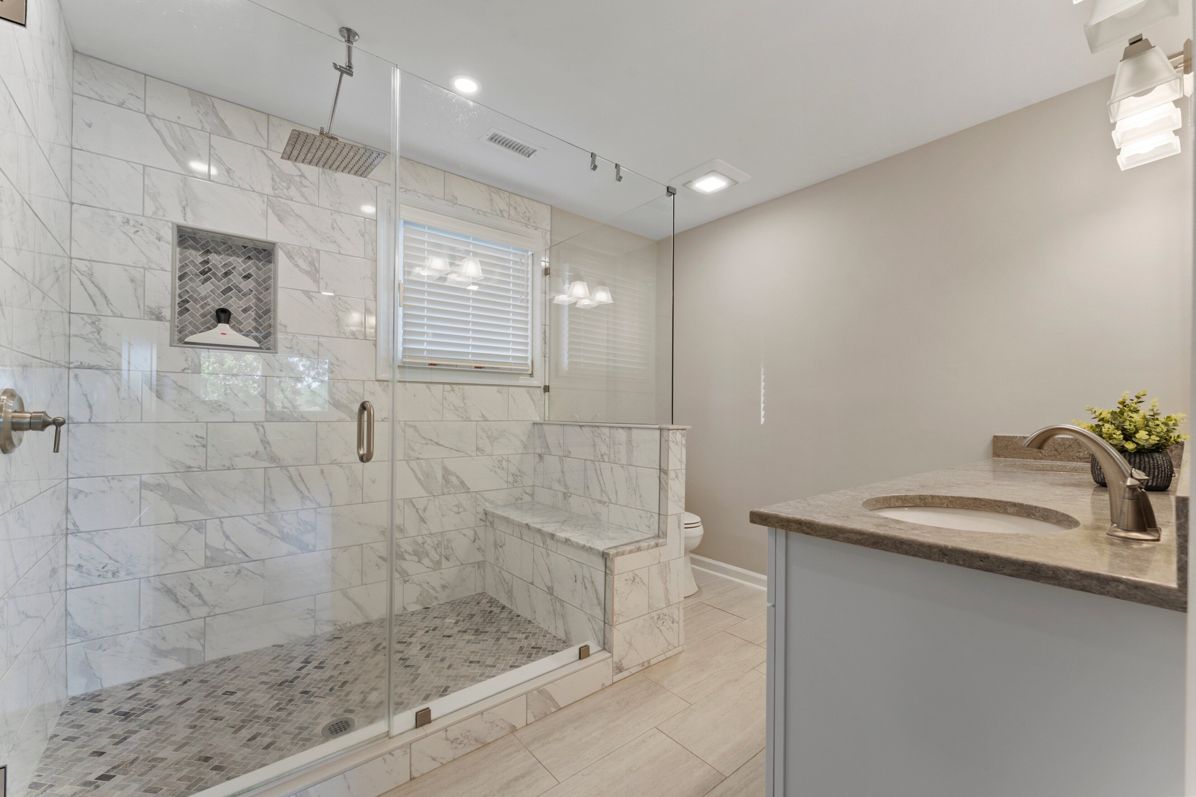 A modern bathroom with a glass-enclosed shower, a marble-tiled wall, a toilet, and a sink with a vase of flowers.