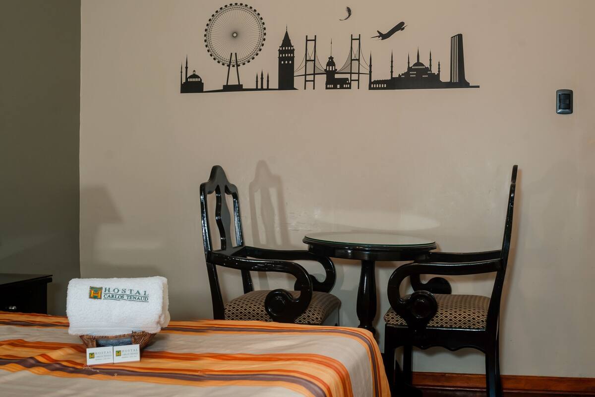 Habitación de hotel con cama, una silla, una mesita y un adorno en la pared que muestra un horizonte urbano con noria y puente.