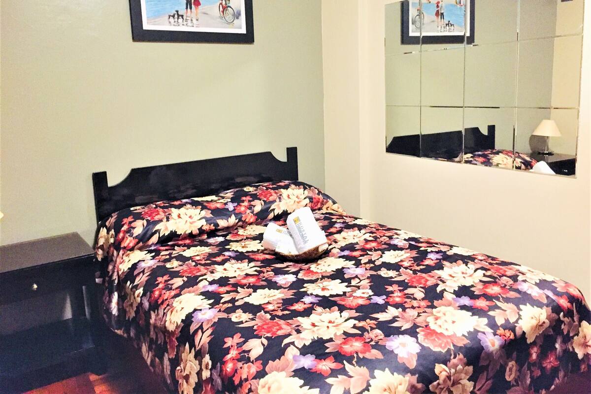 Una cama con colcha estampada, un cuadro enmarcado en la pared y un espejo que refleja la cama y una lámpara.