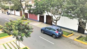A blue car parked on a street with a sidewalk, trees, and a building with a red brick wall and a white fence. - Hostal Carlos Tenaud  (Lima)