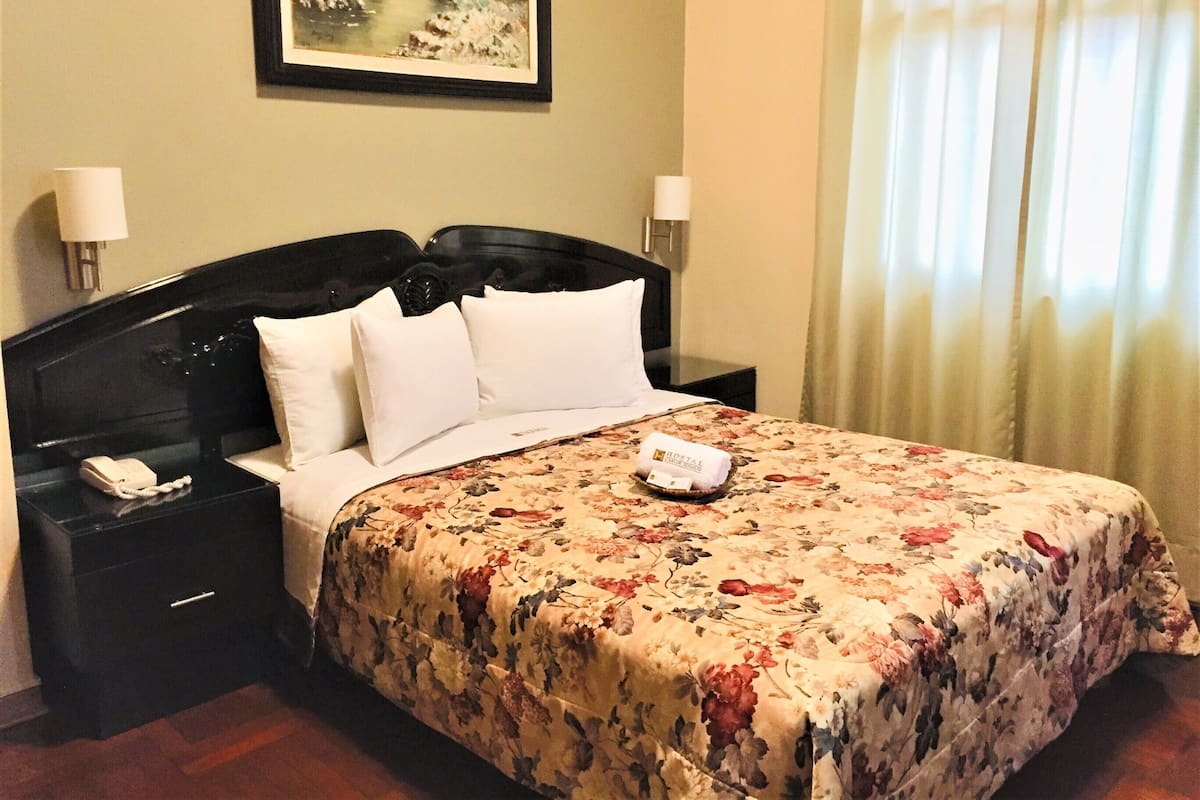 Habitación de hotel con una cama grande, dos mesitas de noche, una lámpara de noche, un cuadro enmarcado en la pared y una ventana con cortinas.