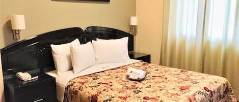 A hotel room with a large bed, two bedside tables, a nightstand, a framed painting on the wall, and a window with curtains.