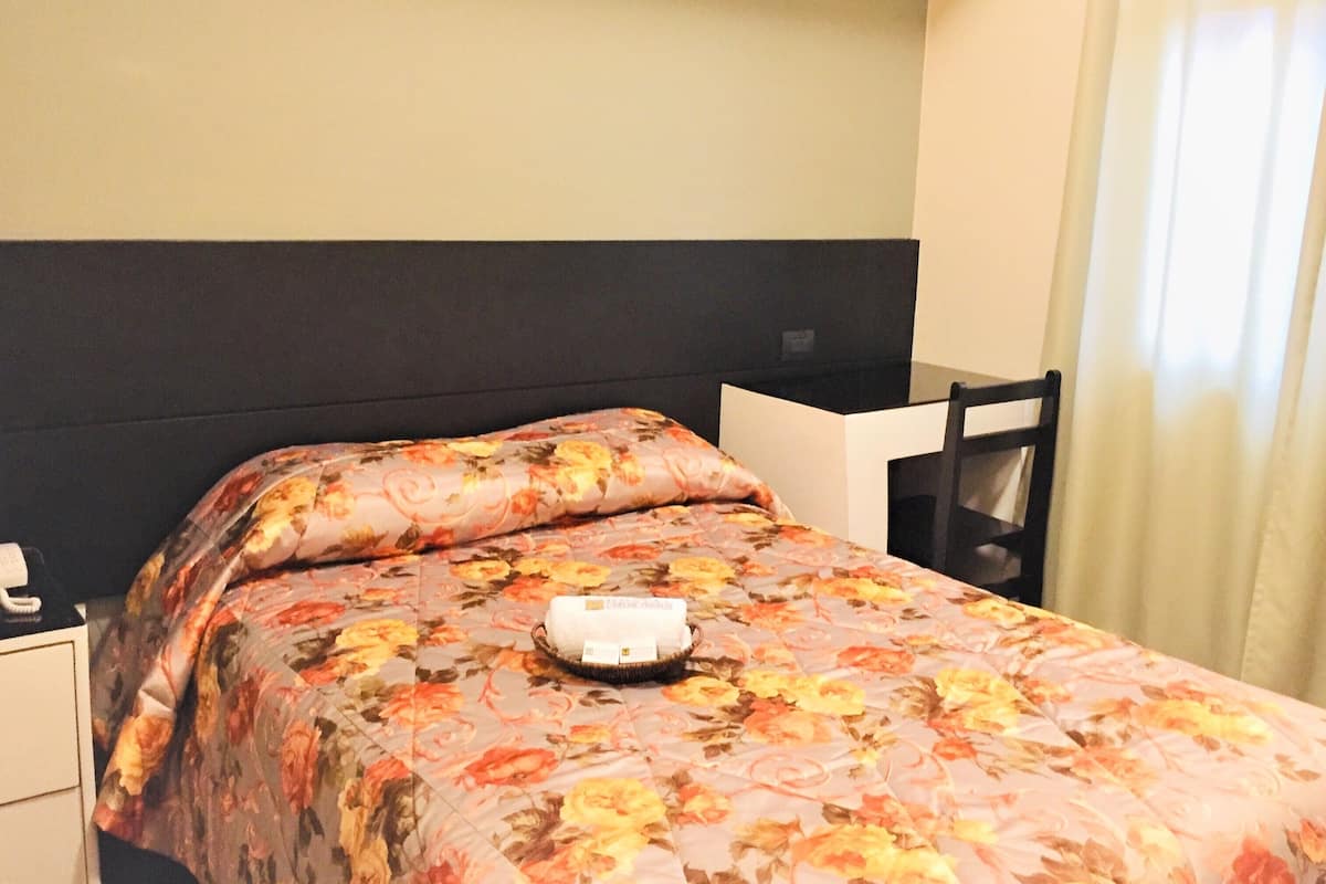 Una cama bien hecha con colcha floral, una mesita de noche con un teléfono y una silla pegada a la pared.