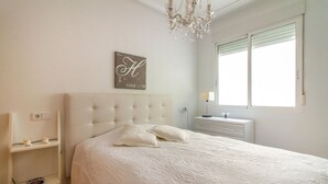 A bedroom with a tufted headboard, a bed with white linens, a bedside table with a lamp, a window with blinds, and a wall-mounted sign. - 2 bedroom amazing apartment in Torrevieja (Torrevieja)